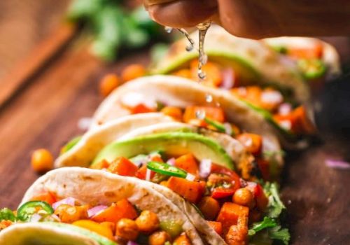 A person's hand squeezes lime juice over a row of chickpea and vegetable tacos, garnished with cilantro and avocado slices, on a wooden board.