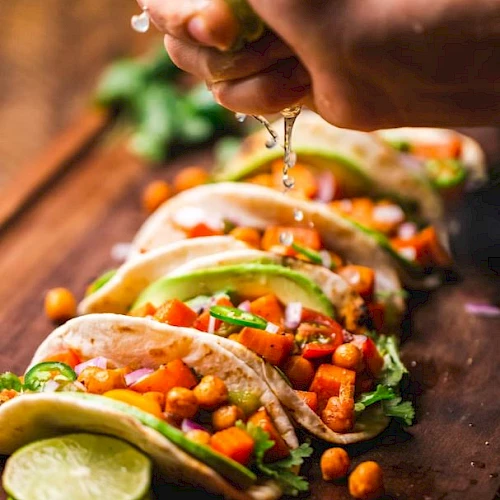 A person's hand squeezes lime juice over a row of chickpea and vegetable tacos, garnished with cilantro and avocado slices, on a wooden board.