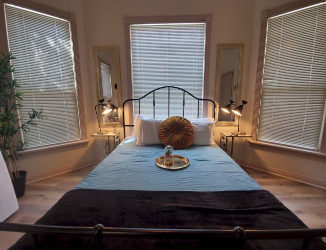 A cozy bedroom with a bed centered between three windows, two mirrors, and bedside tables with lamps; a tray with drinks is on the bed.