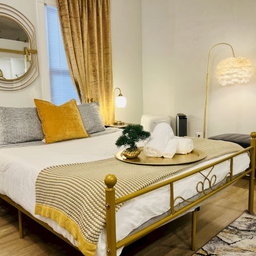 A cozy bedroom with a gold accent bed, decorative pillows, a mirror, a tray with towels, a small plant, warm lighting, and contemporary decor.