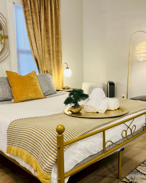 A cozy bedroom with a gold accent bed, decorative pillows, a mirror, a tray with towels, a small plant, warm lighting, and contemporary decor.