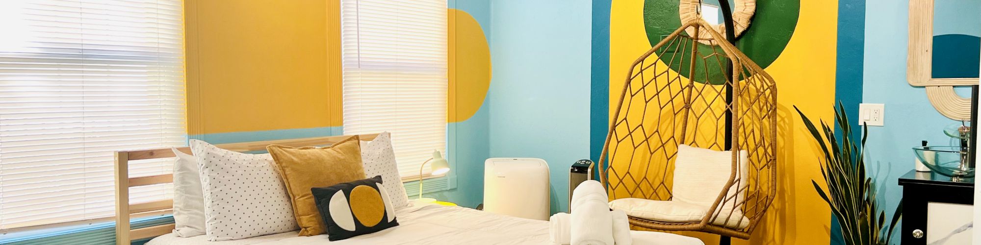 A cozy bedroom with colorful wall art, a wooden bed with white bedding, pillows, a chair, plants, and a rug on the wooden floor.