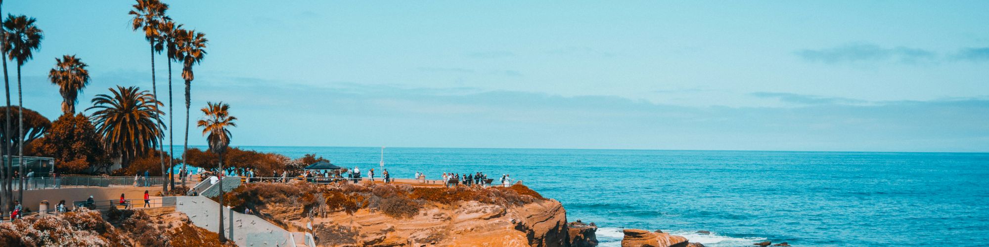 A scenic beach with people enjoying the shoreline, palm trees on the side, and rocks extending into the blue ocean under a clear sky.