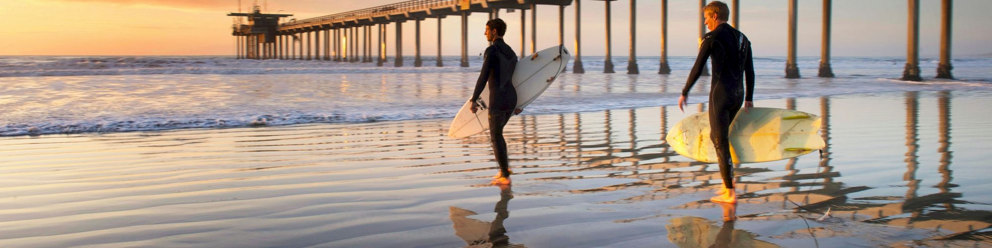 Two surfers walk along the beach, carrying surfboards, at sunset. A long pier extends into the ocean in the background, with calm reflective water.