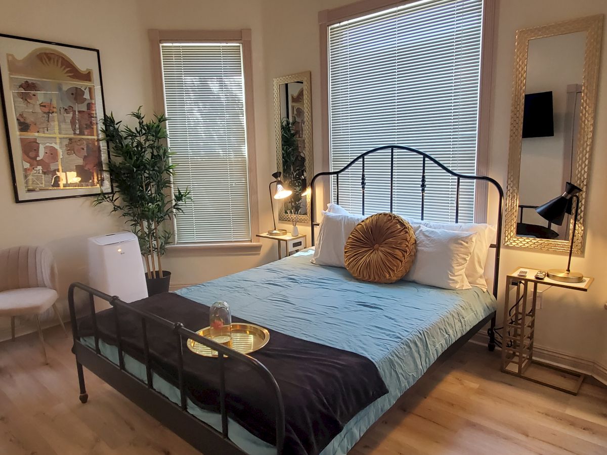 A bedroom with a bed, decorative pillows, a plant, wall art, mirrors, a chair, a white appliance, and a nightstand with lamps and decorative pieces.