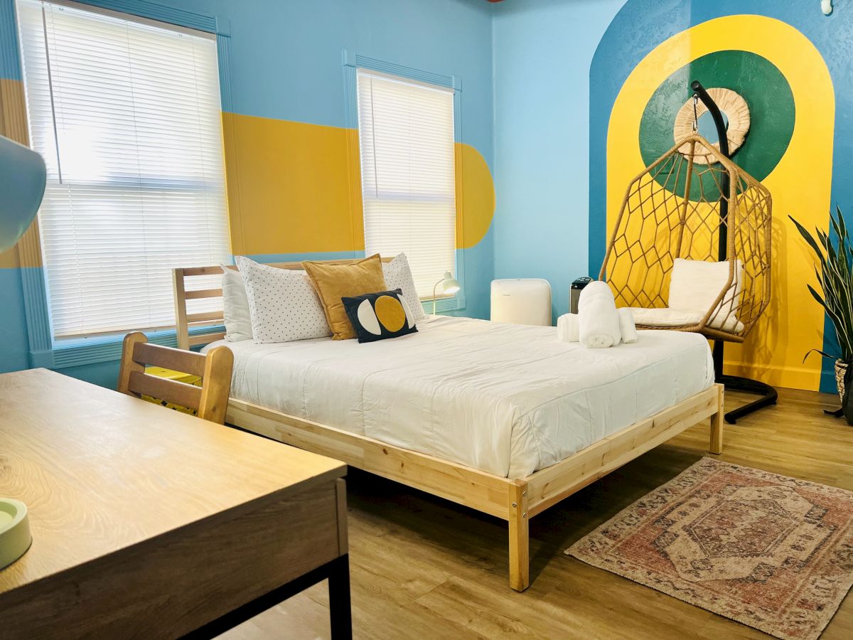 A cozy bedroom with colorful wall art, a bed, hanging chair, desk, and rug. Bright, modern design with a mix of blue, yellow, and natural tones.