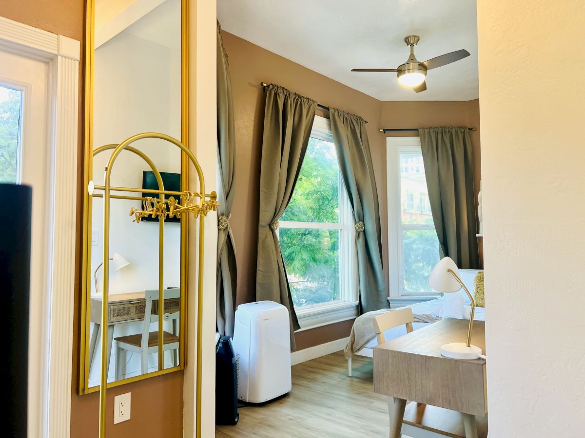 A cozy room with large windows, brown curtains, a ceiling fan, a desk with a lamp, a chair, and a mirror on the wall. There's also a white portable AC unit.