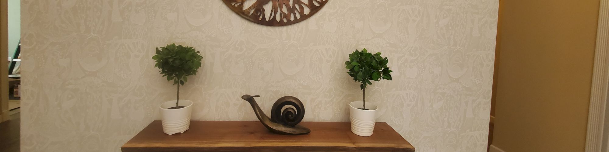 The image shows a wooden table with two potted plants and a decorative snail sculpture, below a circular wall art of a tree.