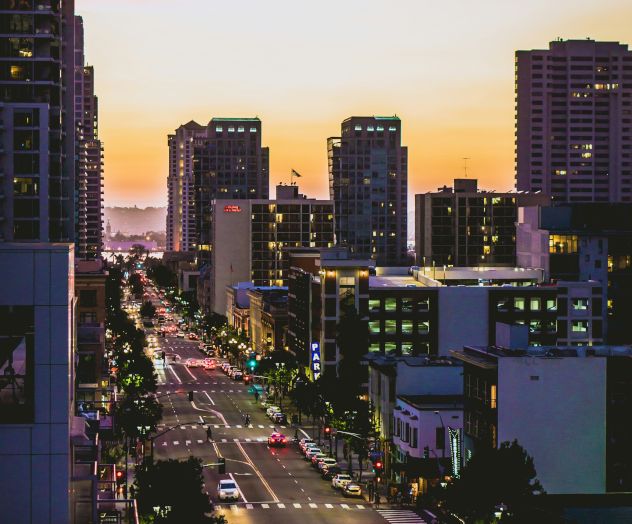 A city scene during sunset, with high-rise buildings, a busy street with cars, and a sky painted with warm colors in the background.