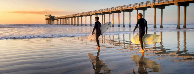 Two surfers holding their boards walk along the wet sand with a pier in the background during sunset.