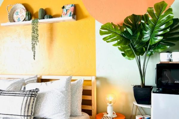 A cozy bedroom features a bed with patterned pillows, vibrant orange wall art, a plant, and a shelf with decor, all creating a bright, cheerful space.