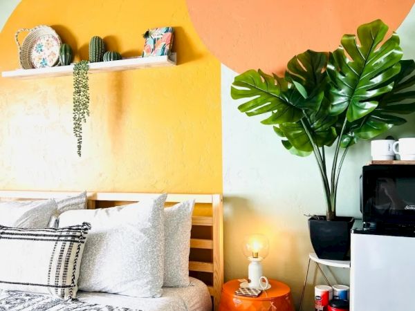 A cozy bedroom features a bed with patterned pillows, vibrant orange wall art, a plant, and a shelf with decor, all creating a bright, cheerful space.