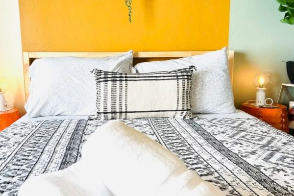 A neatly made bed with a patterned bedspread, pillows, and rolled white towels on top, against a vibrant yellow and green wall with decor.