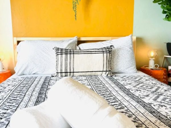 A neatly made bed with a patterned bedspread, pillows, and rolled white towels on top, against a vibrant yellow and green wall with decor.