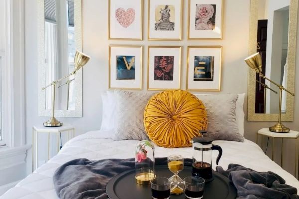 A cozy bedroom with a neatly made bed, a yellow pillow, framed art on the wall, and a tray with drinks on a gray blanket.