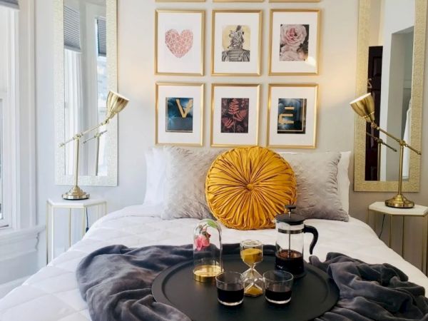 A cozy bedroom with a neatly made bed, a yellow pillow, framed art on the wall, and a tray with drinks on a gray blanket.