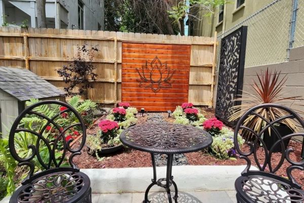 A small garden with ornate metal chairs and table, colorful flowers, and a wooden panel featuring a floral design is shown.