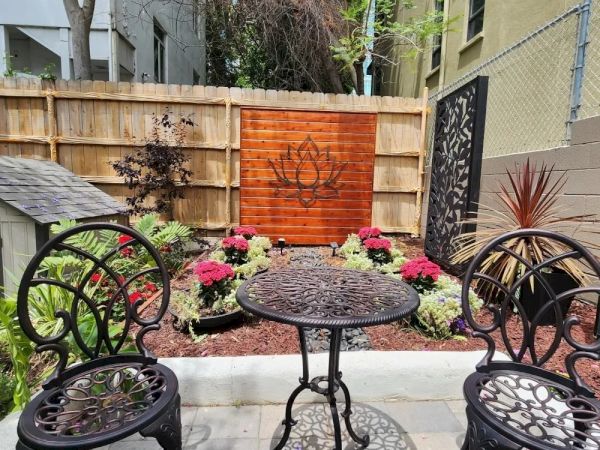 A small garden with ornate metal chairs and table, colorful flowers, and a wooden panel featuring a floral design is shown.