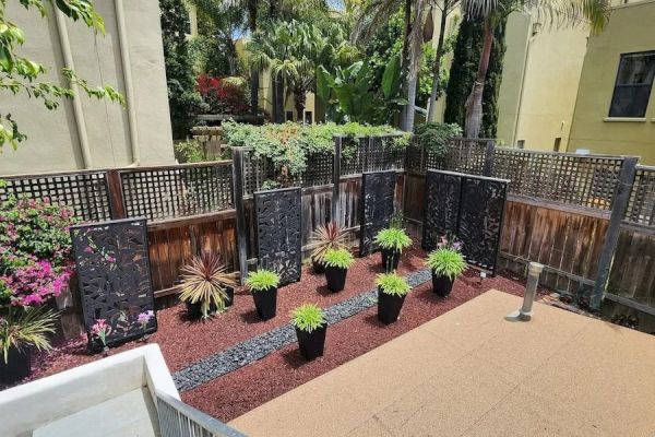 A backyard area with potted plants, decorative screens, palm trees, and a staircase descending to the garden, surrounded by buildings and fencing.