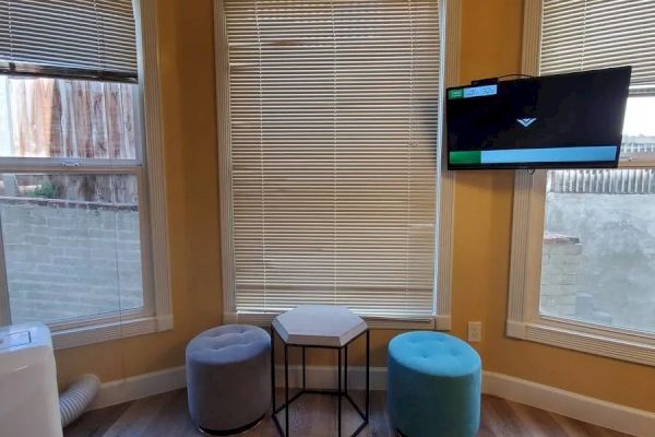 A cozy room corner with three windows, two poufs, a small hexagonal table, and a wall-mounted TV. There’s light streaming in from outside.