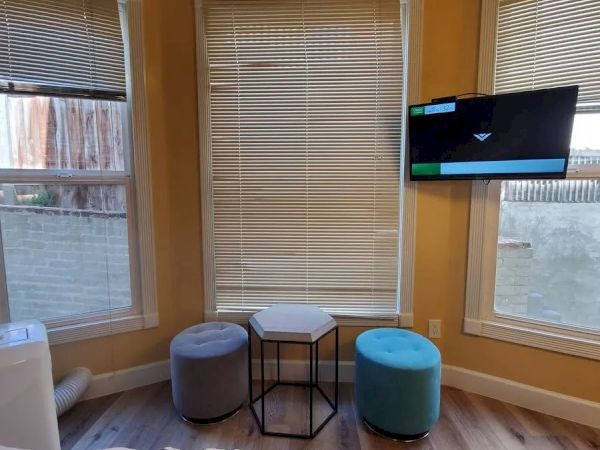 A cozy room corner with three windows, two poufs, a small hexagonal table, and a wall-mounted TV. There’s light streaming in from outside.