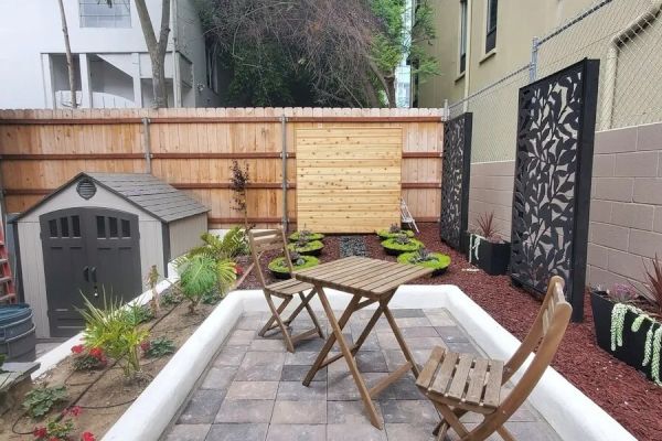 A small, landscaped backyard with a wooden table and chairs, potted plants, a garden shed, and decorative panels on the walls near the fence.