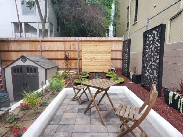 A small, landscaped backyard with a wooden table and chairs, potted plants, a garden shed, and decorative panels on the walls near the fence.