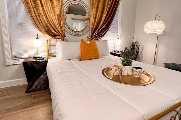 A cozy bedroom with a neatly made bed, gold and white decor, a tray with mugs and a plant on the bed, and elegant curtains.