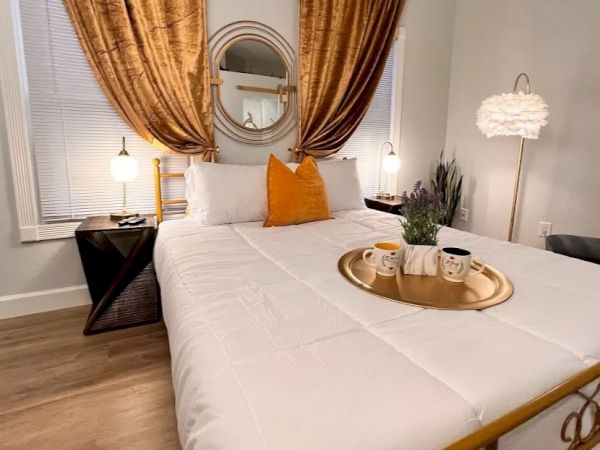 A cozy bedroom with a neatly made bed, gold and white decor, a tray with mugs and a plant on the bed, and elegant curtains.