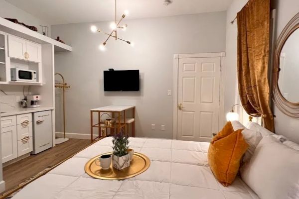 This image shows a modern, cozy bedroom with a neatly made bed, wall-mounted TV, a kitchenette, and tasteful decor including a circular mirror and plant.