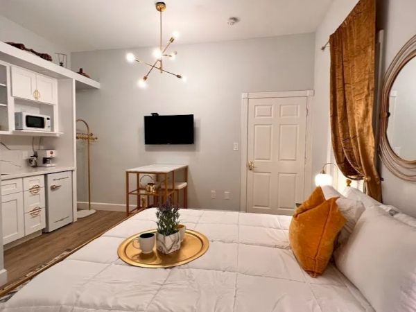 This image shows a modern, cozy bedroom with a neatly made bed, wall-mounted TV, a kitchenette, and tasteful decor including a circular mirror and plant.