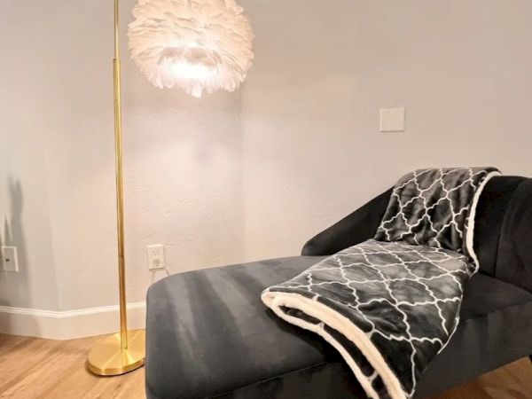 The image shows a cozy corner with a black chaise lounger, a geometric-patterned blanket, wood flooring, and a gold floor lamp with a feathery shade.