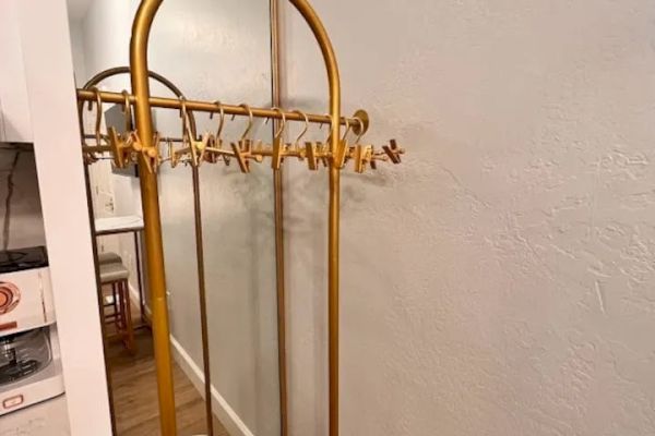 The image shows a yellow coat rack with multiple hangers in a hallway next to a large mirror. Part of a kitchen area is visible to the left.