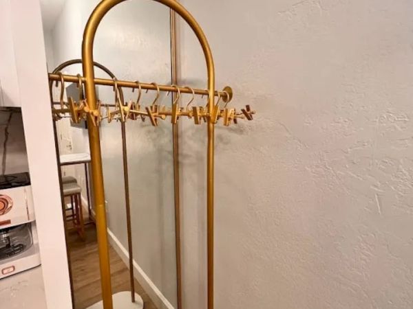 The image shows a yellow coat rack with multiple hangers in a hallway next to a large mirror. Part of a kitchen area is visible to the left.