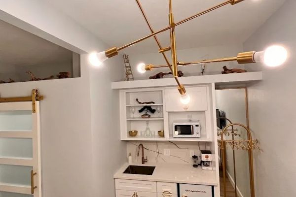 A small kitchenette with white cabinets, a sink, a microwave, and a modern brass light fixture. There's a sliding door and a coat rack.