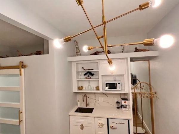 A small kitchenette with white cabinets, a sink, a microwave, and a modern brass light fixture. There's a sliding door and a coat rack.