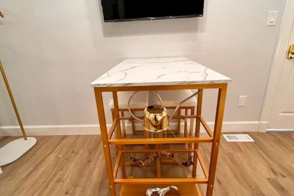 The image shows a room with a wall-mounted TV above a small wooden shelving unit with a white top and gold accents, placed on wooden flooring.