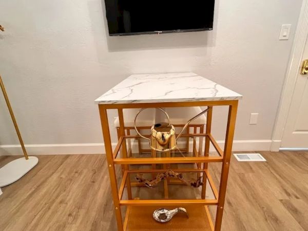 The image shows a room with a wall-mounted TV above a small wooden shelving unit with a white top and gold accents, placed on wooden flooring.