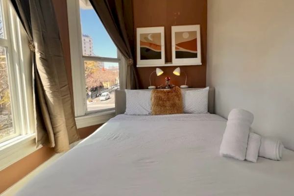 A cozy bedroom with a double bed, white linens, two framed pictures above the headboard, and a window with drapes letting in natural light.