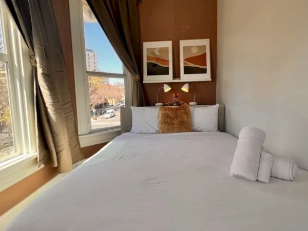 A cozy bedroom with a double bed, white linens, two framed pictures above the headboard, and a window with drapes letting in natural light.