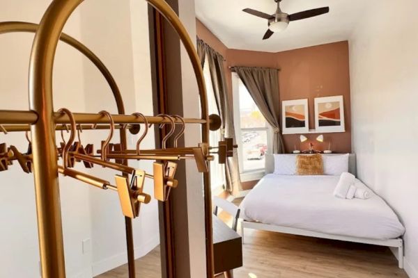 The image shows a minimalistic bedroom with a neatly made bed, wall art, a ceiling fan, wooden flooring, and a golden luggage rack in the foreground.