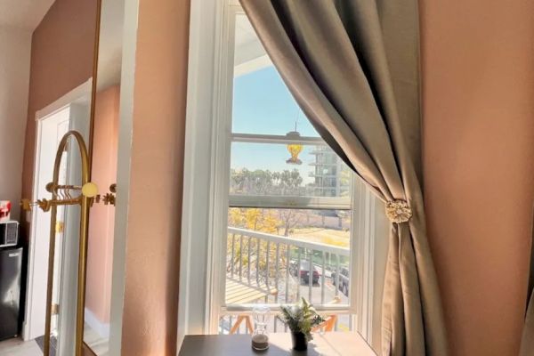 A desk with a plant is positioned by a window with taupe curtains, overlooking a sunny exterior scene, featuring a road and greenery outside.