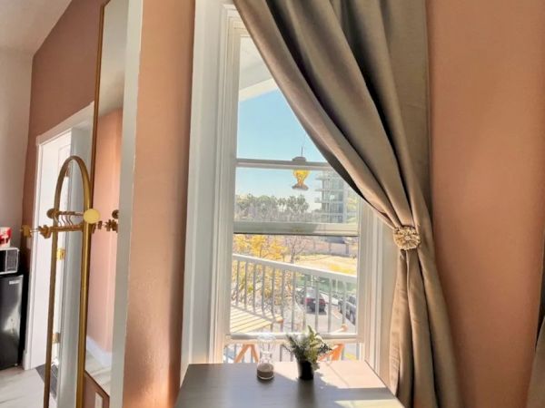A desk with a plant is positioned by a window with taupe curtains, overlooking a sunny exterior scene, featuring a road and greenery outside.