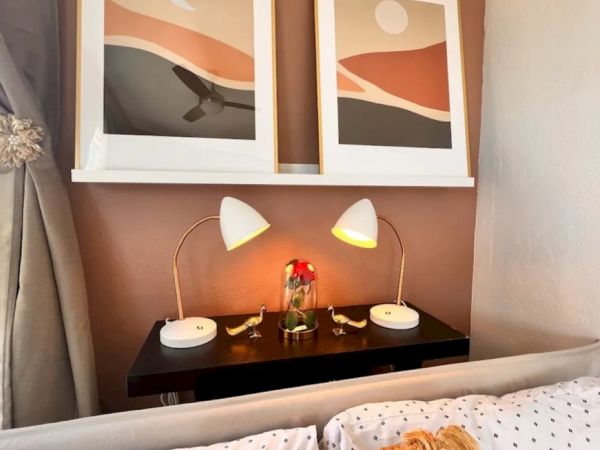A cozy bedroom setup with two abstract art pieces, matching bedside lamps, a vase with flowers, a cushioned bed, and polka-dot bed sheets.