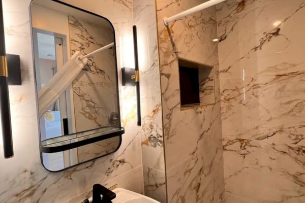 A modern bathroom with marble walls, a rectangular mirror with black frame, wall lights, a sink with a black faucet, and a corner shower area.