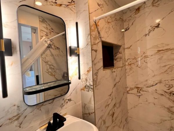 A modern bathroom with marble walls, a rectangular mirror with black frame, wall lights, a sink with a black faucet, and a corner shower area.