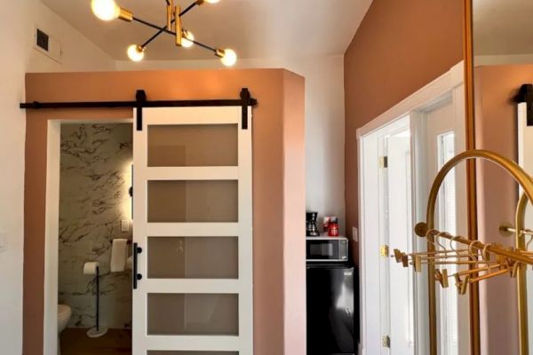 A modern interior with a stylish chandelier, pink walls, a sliding door to a bathroom, a coat rack, and a small kitchenette with a microwave and coffee maker.