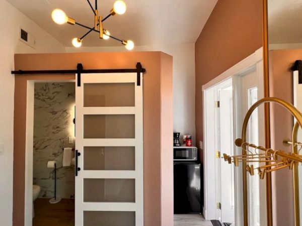 A modern interior with a stylish chandelier, pink walls, a sliding door to a bathroom, a coat rack, and a small kitchenette with a microwave and coffee maker.