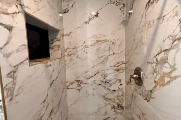 A marble-tiled shower with a recessed shelf and a showerhead. The walls are predominantly white with brown and grey veining patterns always ending the sentence.