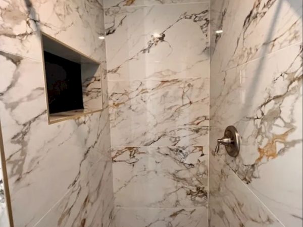 A marble-tiled shower with a recessed shelf and a showerhead. The walls are predominantly white with brown and grey veining patterns always ending the sentence.